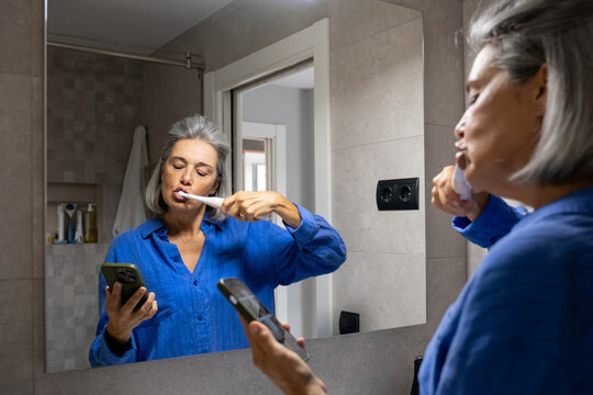 Mature woman brushes teeth and checks phone in bathroom