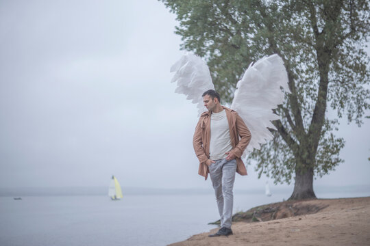 Elegant man with white angel wings walking on natural lakeside beach with copy space