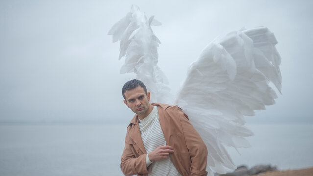 Serious man with large white angel wings looking at camera on misty waterfront with copy space