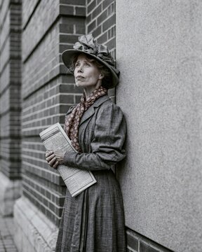 1900s suffragette holding a pamphlet beside a large blank brick building facade