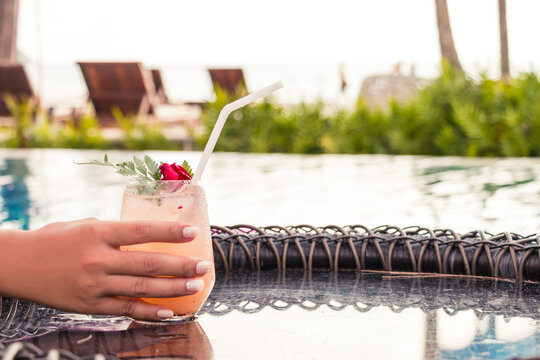 Hand of woman holding lychee soda cocktails on table with blurred poolside and beach background. Summer drinks concept.