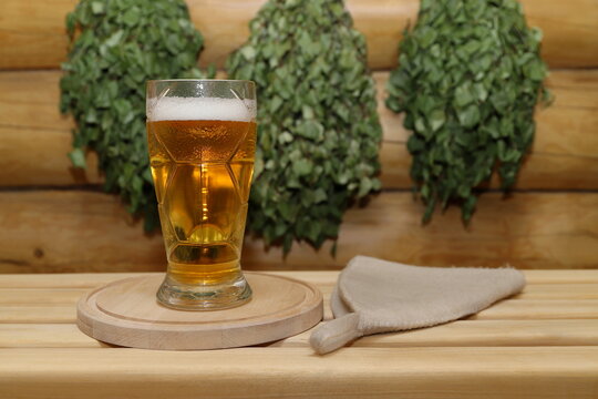 On a plank bench, next to a sauna hat, stands a glass goblet of light beer on a wooden round tray, against the background of log wall with a birch brooms.