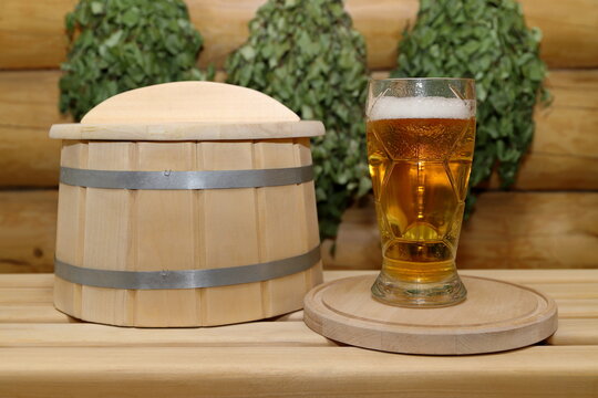 On a plank bench, next to a wooden barrel, stands a glass goblet of light beer on a wooden round tray, against the background of log wall with a birch brooms. 
