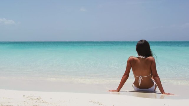Travel vacation girl in white bikini relaxing in tropical beach sun tanning in summertime.W oman in relaxation mode at idyllic stunning tropical luxury resort hotel. SLOW MOTION.