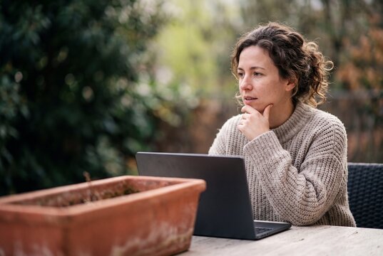 Thoughtful woman working remotely on laptop outdoors at garden table, contemplating ideas in peaceful backyard setting