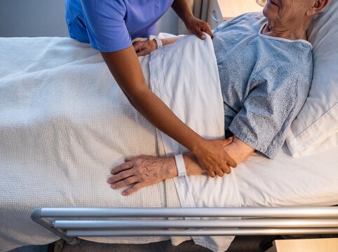 Nurse adjusting a blanket for an elderly male patient lying in a hospital bed, providing compassionate care and support