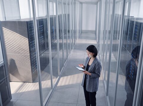 Asian female IT engineer using a digital tablet while standing in a modern corporate data center corridor