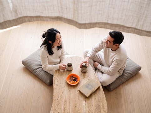 Happy young multiethnic couple sitting on floor cushions drinking coffee and chatting at home