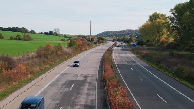 Curving dual carriageway through rural valley with passenger cars and white truck moving away from elevated roadside viewpoint; motion blur on nearest vehicles suggests steady forward travel under