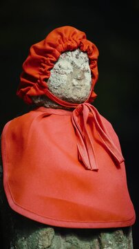 cinematic vertical close-up shot of an old stone jizo statue wearing a red bib and cap against a dark background at a japanese temple