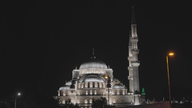Slow motion 2x 4K footage of a beautifully lit mosque with a central dome and a tall minaret standing in the dark night sky with warm street lamps nearby in Istanbul