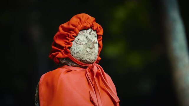cinematic horizontal shot of an old stone jizo statue wearing a red bib and cap against a dark moody background at a japanese temple panning right