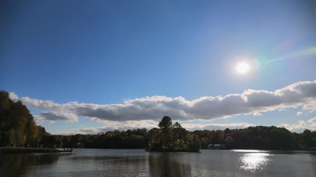Timelapse Trees with yellow and orange leaves grow along the edge of a river. The sky is clear with no clouds. The scene shows the change of seasons in a park.