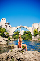 female tourist with backpack sitting on rock looking at stari most bridge in mostar bosnia and...