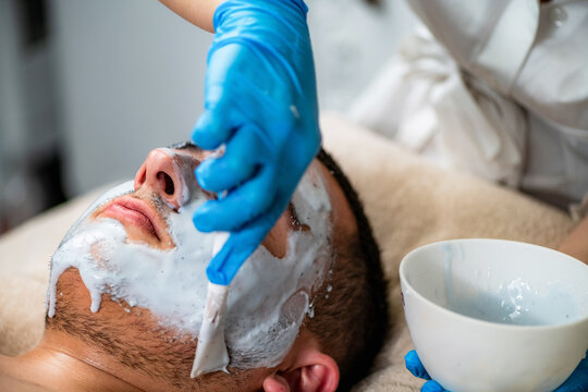 Acne treatment for a young man receiving professional cosmetic facial care in beauty salon, with esthetician applying a skincare mask for cleansing and blemish care.