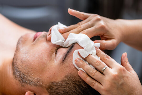Acne treatment for a young man receiving professional facial cleansing in beauty salon, with esthetician removing cosmetic product and impurities from the skin.