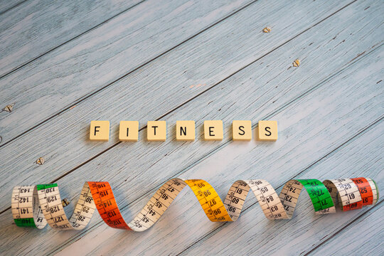 Top-down composition on a vintage blue wooden surface showing the word "FITNESS" formed with board game pieces. Below the letters, a rolled-up measuring tape extends, representing tracking physical pr