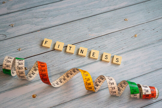Conceptual composition on a weathered wooden surface, with letter tiles forming the word fitness and a colorful measuring tape. Concept of exercise, physical activity, self-care, and health.