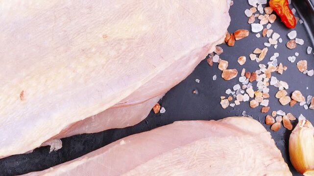 Raw chicken breast placed on a black plate alongside coarse salt crystals and a red chili pepper. A two-pronged carving fork and a black knife rest beside the plate on a dark textured surface,.