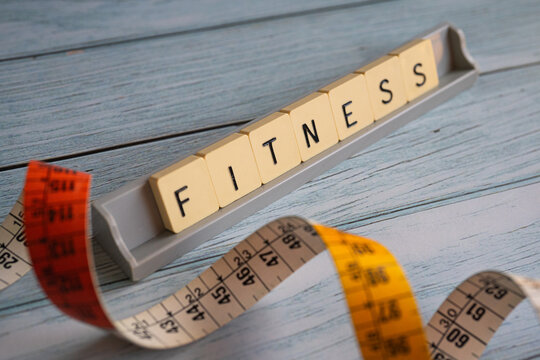 Conceptual image related to fitness, health, and wellness. A measuring tape surrounds letter tiles spelling "FITNESS" on a blue wooden surface. Perfect for illustrating exercise, weight control, train