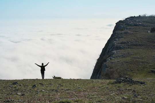 Woman silhouette with raised arms stands on mountain peak edge looking at white cumulus cloud sea surface below daylight sky. Spacious aerial outdoor scene