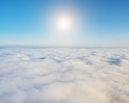 Bright sunburst shines in blue sky above dense white cumulus clouds sea surface at high altitude daylight. Spacious aerial lamdscape.
