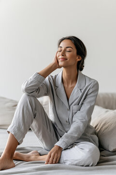 Serene young woman relaxing peacefully on a bed while wearing comfortable grey striped pajamas in a bright minimalist bedroom.