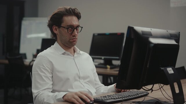 Tired young businessman in glasses working on computer in modern office. Exhausted, overworked employee feels sleepy, bored and yawns at desk. Burnout and fatigue concept. Shooting in slow motion.