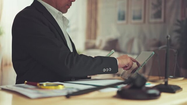 Mature asian lawyer reviewing digital documents on tablet computer beside gavel, scale justice, paperwork on office desk.