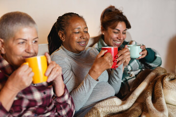 Plakat Happy multiracial senior women drinking hot tea together while sitting on couch during winter time - Cozy, elderly women friendship and lifestyle concept