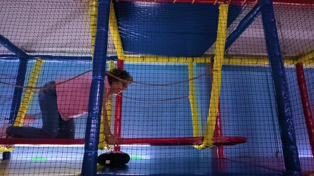 A joyful mature Latina woman crawling across a swinging red platform inside a colorful indoor children's playground obstacle course in Mexico City.