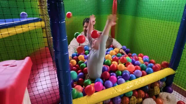 A high-angle shot of a latino father and his toddler son throwing plastic balls and laughing while playing inside a colorful indoor playground ball pit.