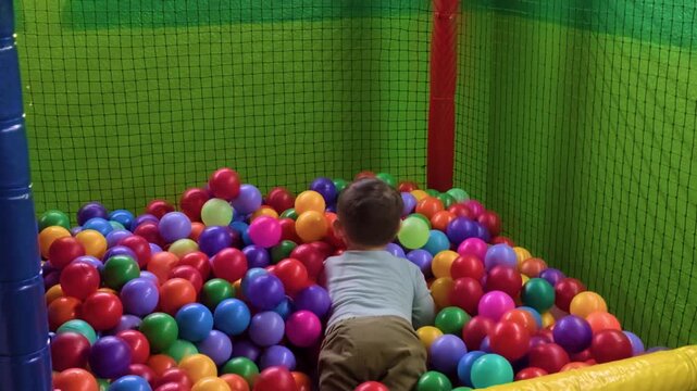 A happy Mexican Latino toddler plays and laughs while diving into a vibrant multicolored ball pit inside a Ninja obstacle course area for children.