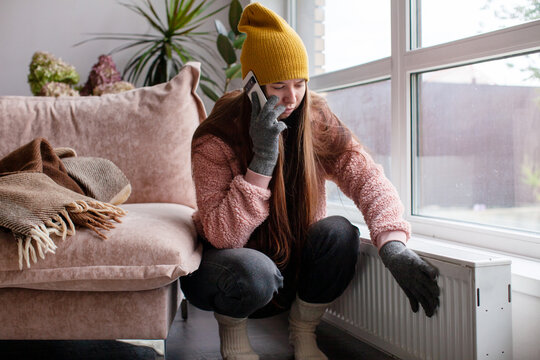 Woman checking cold radiator at home while wearing winter clothes. No heating or broken heating concept. Household energy poverty and rising utility costs. Winter comfort and home