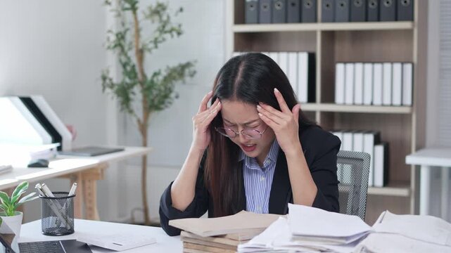 Stressed asian businesswoman experiencing headache and burnout from overworking with a pile of paperwork in the office