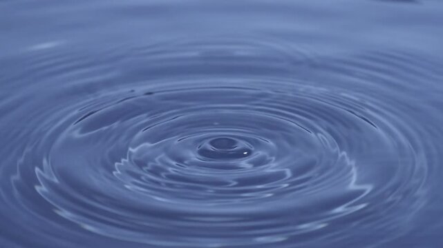 Single water drop falling into blue liquid surface creating a crown