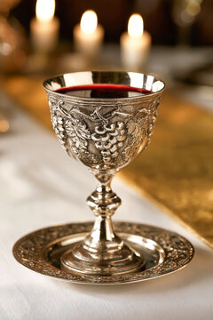 A silver Kiddush cup sits on a saucer, filled with red wine. The setting features a white tablecloth, warm candlelight, and a golden table runner in