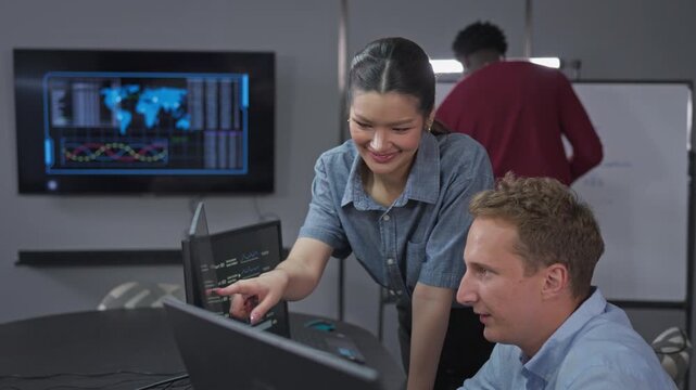 Woman leans over a desk and points at charts on a coworker's screen while he talks through the numbers, with teammates at a whiteboard behind.