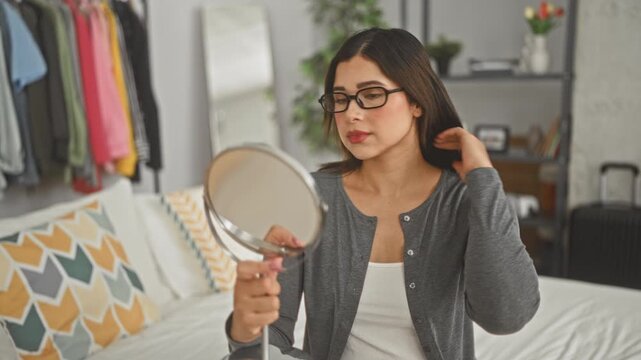 Woman adjusts glasses while holding a handheld mirror on a bed inside a building bedroom, looking into her reflection with calm focus; introspection serenity.