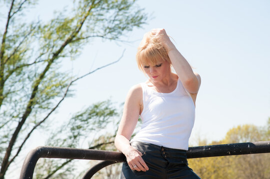 Reife blonde Frau mit Pony posiert selbstbewusst outdoor in wei&szlig;em Top, schwarzer Lederhose und hohen Stiefeln. Best Ager, Casual Fashion, erfahrene Lady, Lifestyle, Sommer und moderne Outdoor-Fotogra