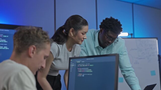 Woman and Black colleague in glasses lean in to read code on a monitor as she explains a change, while a teammate watches from his seat.