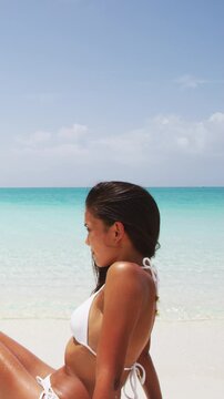 Vacation on beach - woman sitting in edge of water enjoying sunbathing on Caribbean white sand paradise beach. Vertical video of real unretouched woman model without any makeup