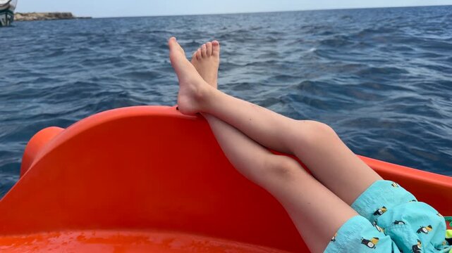 Boy rests on orange boat with legs crossed, relaxing beside blue sea. Child in patterned swimwear enjoys quiet catamaran ride while small waves ripple across open water, resting aboard boat.