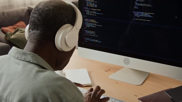 African American mature software engineer wearing headphones typing code on computer. Footage suitable for remote work campaigns, coding bootcamps, and IT technology marketing