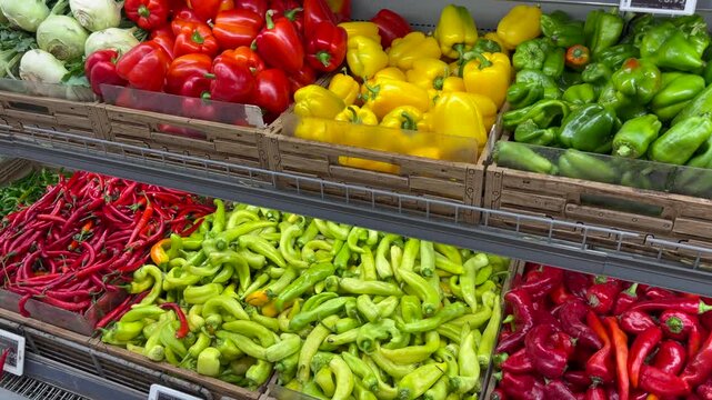 Fresh bell peppers and hot peppers are arranged in wooden market bins. Red, yellow, and green vegetables form colorful grocery display, with abundant produce ready for shoppers to select.