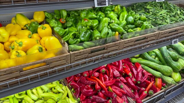 Rows of colorful peppers and chilies occupy wooden produce crates at grocery store. Green, red, and yellow vegetables are arranged by variety beneath shelf price labels, separating sweet peppers