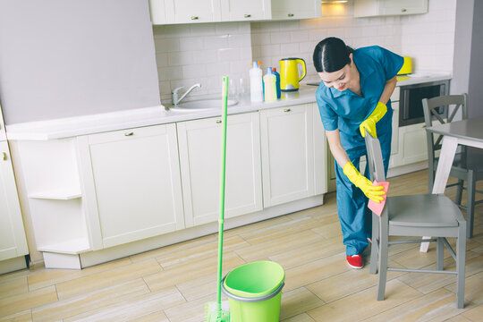 Concentrated and hard-working girl works in kitchen. She looks down and cleaning chair with orange rag. There are green bucket and mop on floor.