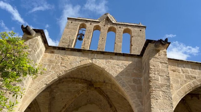 Kyrenia, Cyprus - October 3, 2026 Bellapais Monastery church tower rises above sunlit stone arches, with bell framed by open windows. Blue sky and leafy branches enrich this historic landmark.