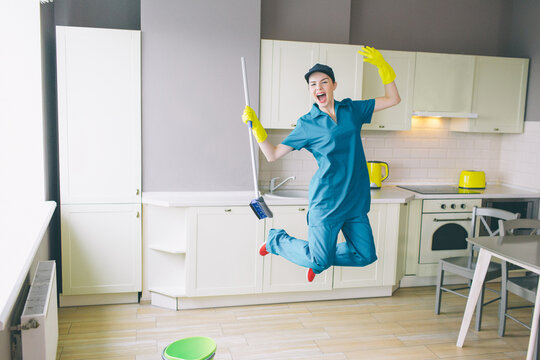 Funny cleaner jumps up in kitchen. Whe keeps her hands up. Girl has rest. She stopped cleaning. Woman looks on camera. She is happy.