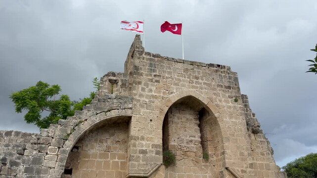Kyrenia, Cyprus - October 3, 2026 Bellapais Abbey rises beneath overcast skies, its aged Gothic masonry and arched gateway framed by trees while Turkish and Northern Cyprus flags wave overhead.